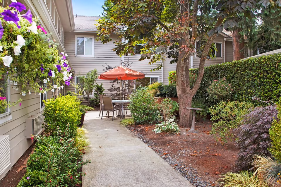 A landscaped courtyard with a concrete walkway leading to a patio table and red umbrella surrounded by flowering plants and the building exterior.