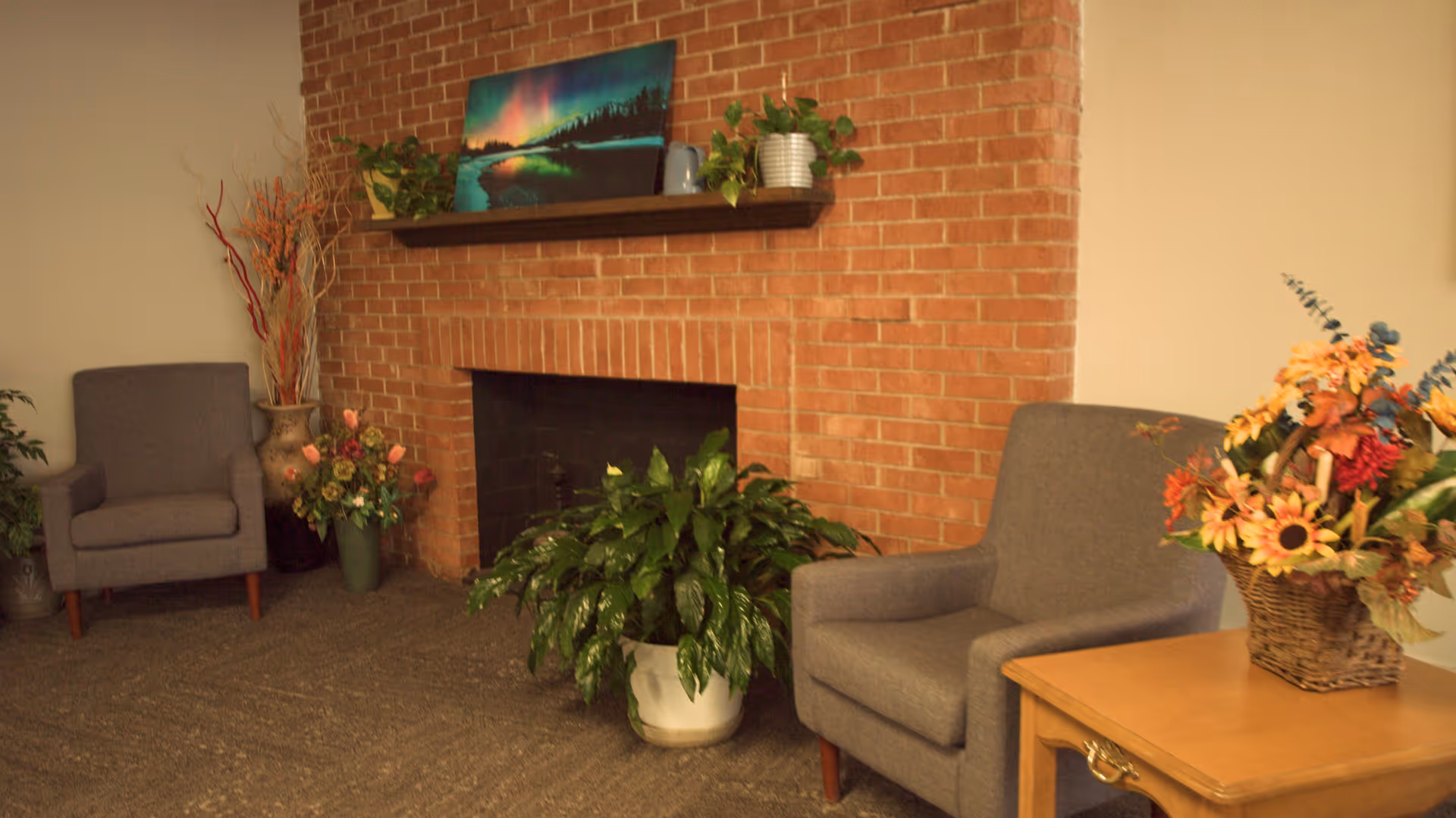 A cozy sitting area with two gray armchairs placed on either side of a brick fireplace. The fireplace has a wooden mantel with a landscape painting and two potted plants. There are additional plants and floral arrangements around the fireplace and on a wooden side table next to one of the chairs.