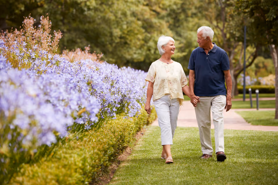 An elderly couple holding hands and walking along a grassy path next to a flower bed with purple flowers in a park-like outdoor setting with trees in the background.