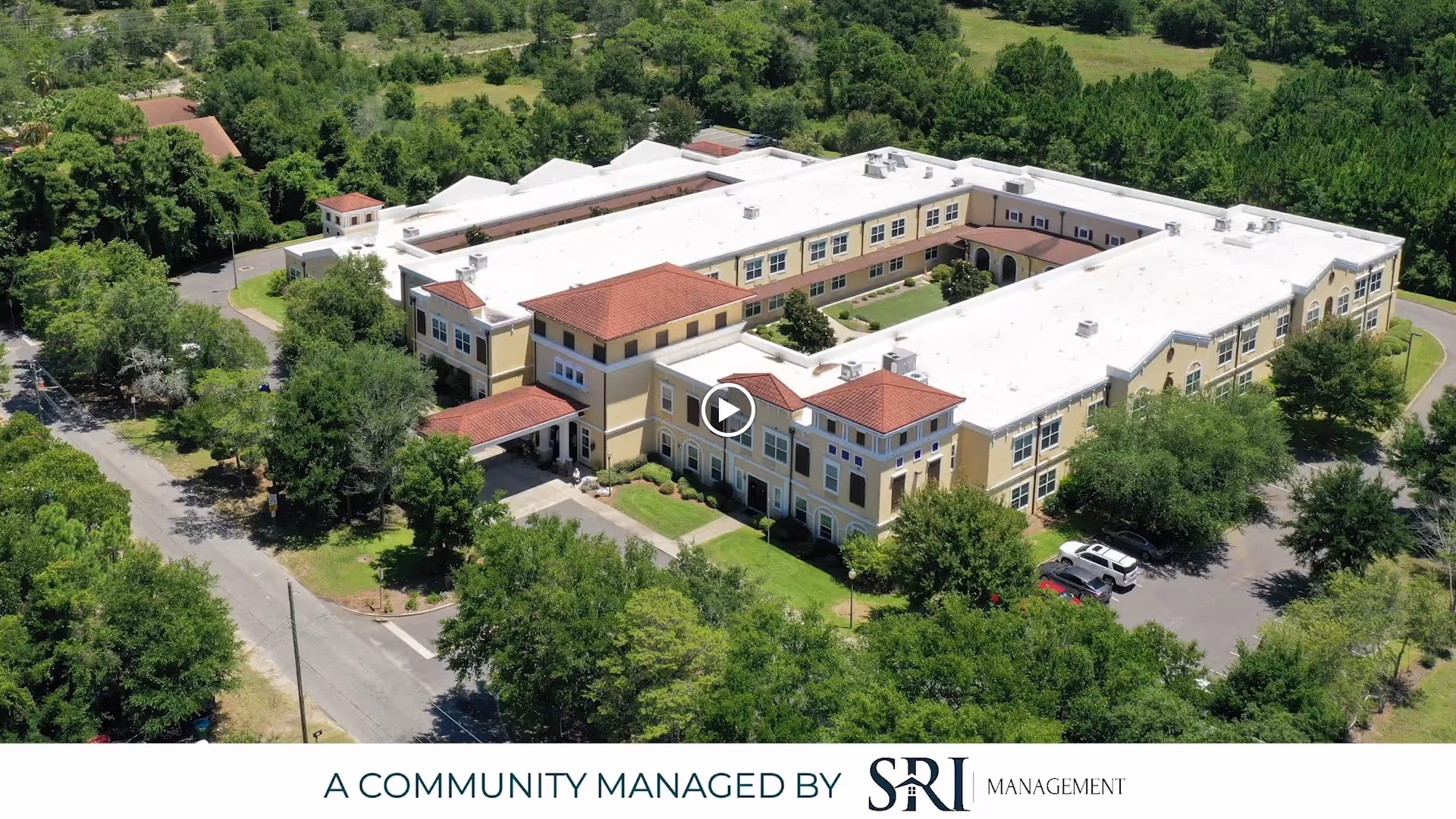 Aerial view of The Beacon at Gulf Breeze, a large senior living facility surrounded by trees and greenery, with a parking area and roads nearby.