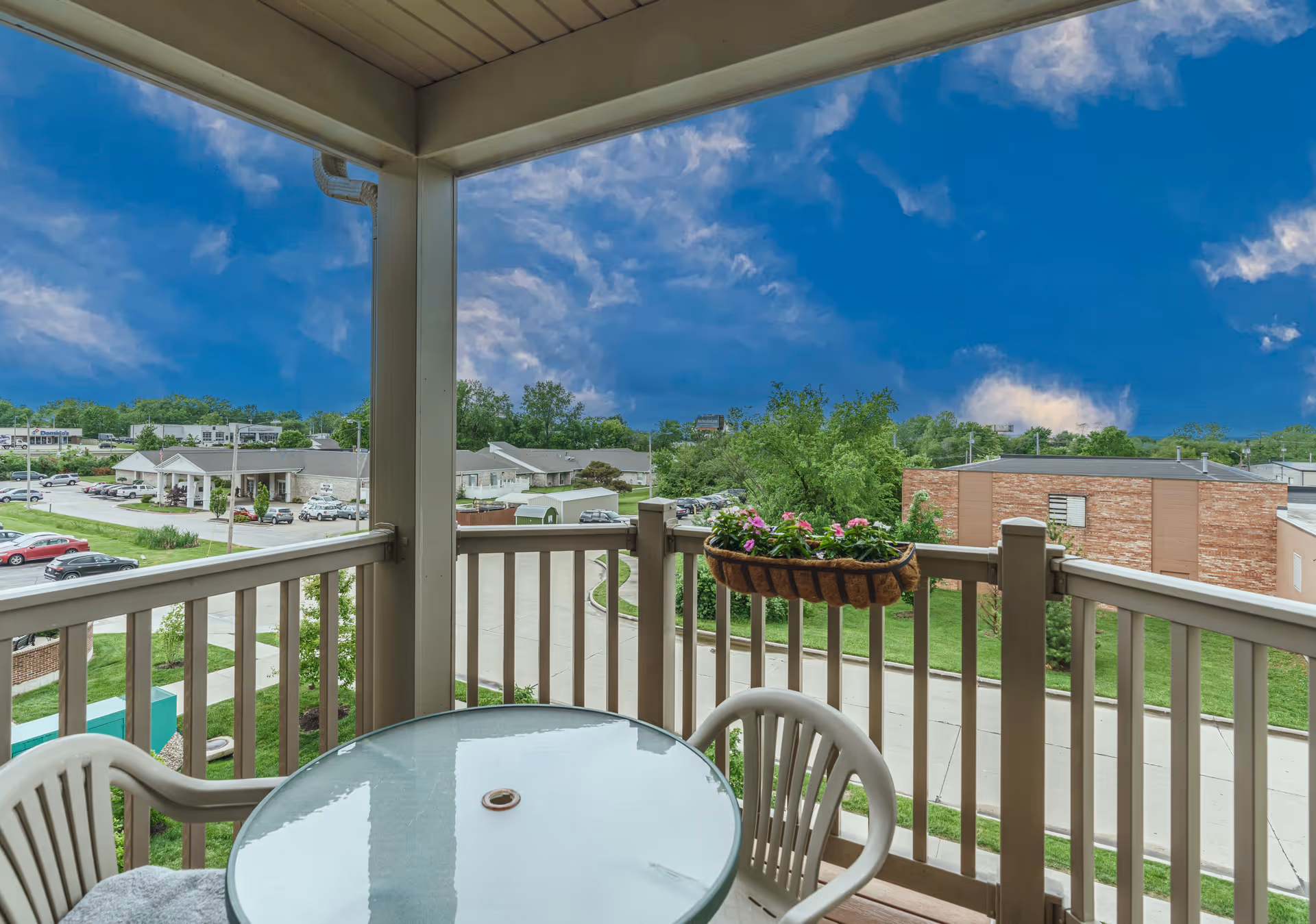 View from a covered balcony with a round glass table and plastic chairs, overlooking a parking lot, buildings, trees, and a partly cloudy blue sky.