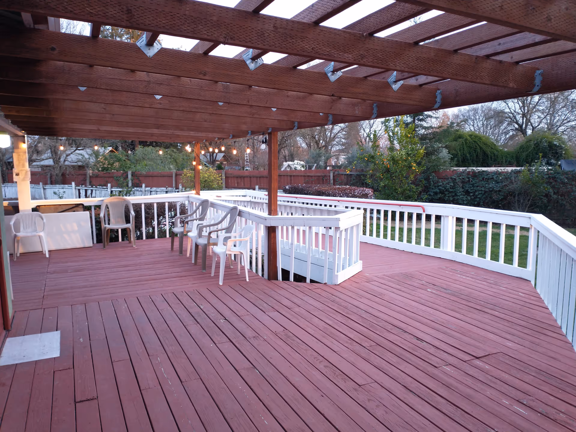 Outdoor wooden deck area with a pergola overhead, several plastic chairs arranged along the railing, string lights hanging from the pergola, and a grassy yard with trees and bushes in the background.