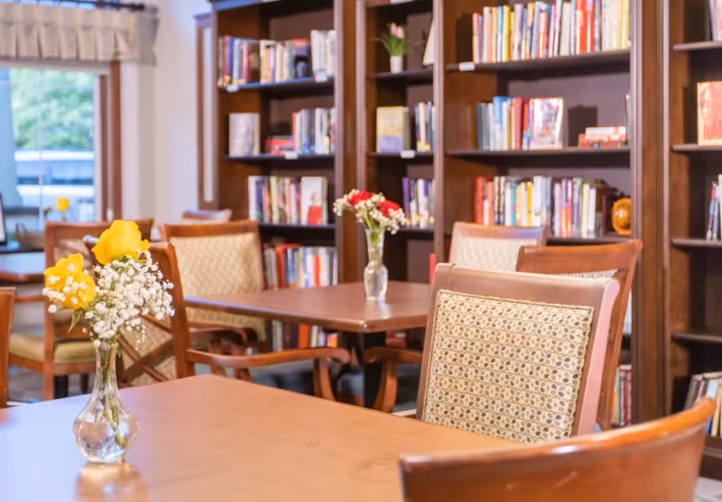 A cozy library or reading room with wooden tables and chairs featuring patterned cushions. There are small vases with fresh flowers on the tables. In the background, there are tall wooden bookshelves filled with books. A window with a light curtain allows natural light into the room.