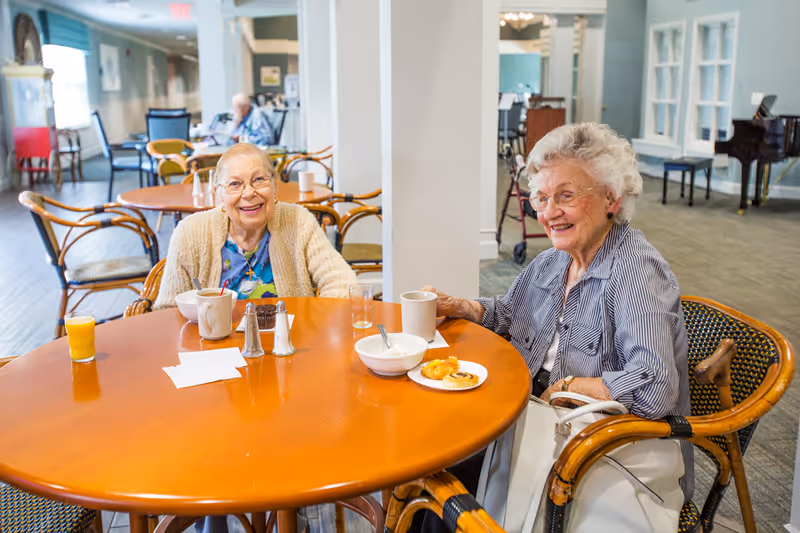 Two elderly women sitting at a round wooden table in a dining area, smiling and enjoying a meal with coffee, juice, and pastries. The background shows more tables and chairs, a piano, and a bright, spacious room.