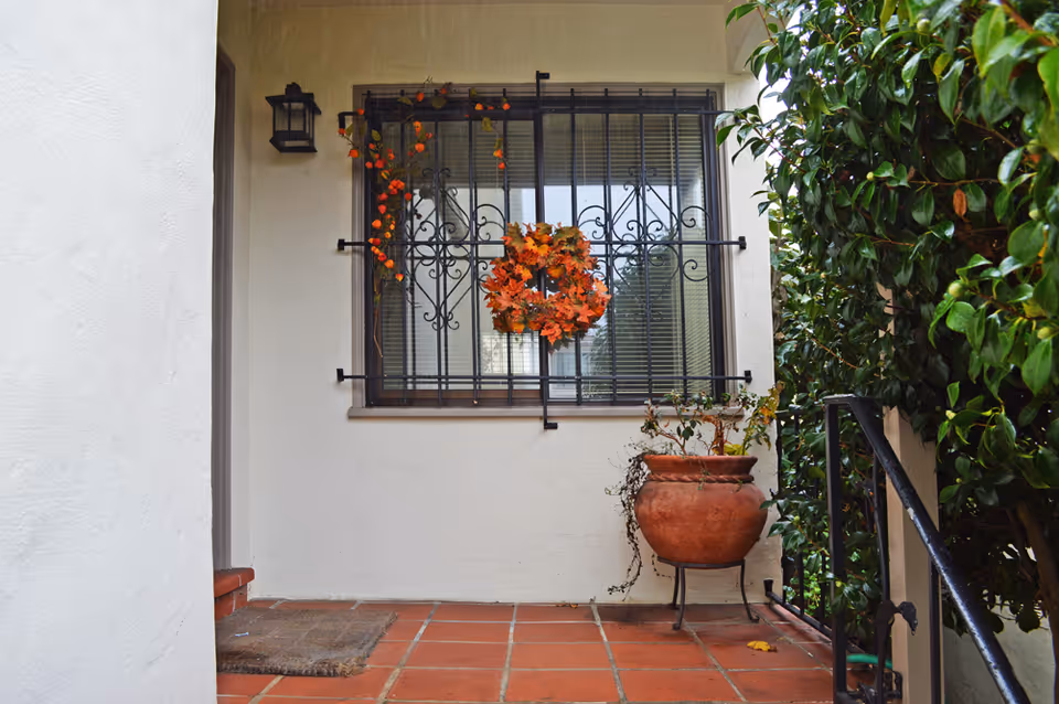 A small outdoor entryway with terracotta tiled floor, a window with decorative black iron bars adorned with an autumn-themed wreath and orange leaves, a large terracotta pot with a plant on a metal stand, and green foliage on the right side.