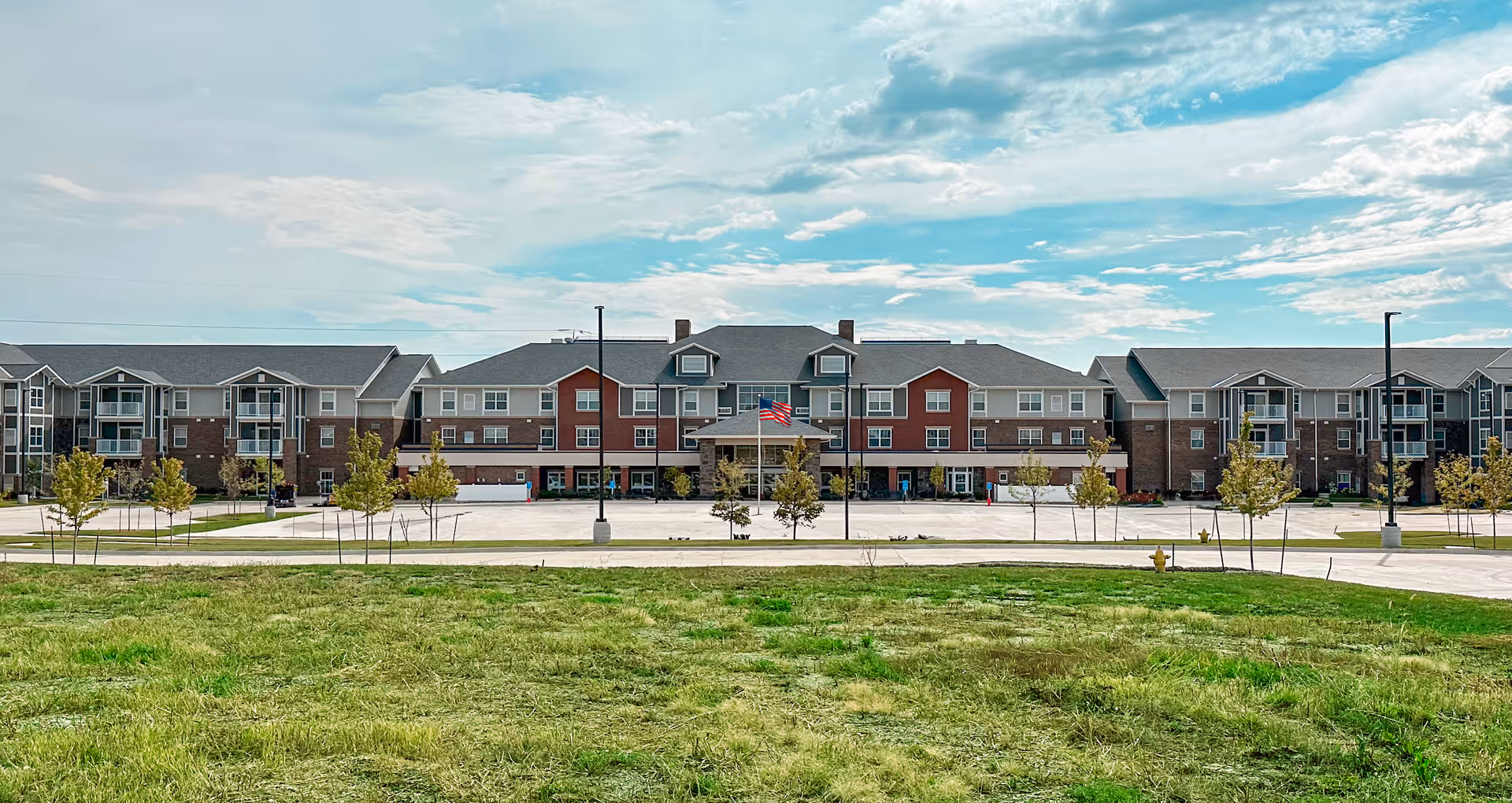 Front exterior of the Glen Meadows senior living building — a three-story complex with a central entrance and American flag, parking area, and grassy foreground.