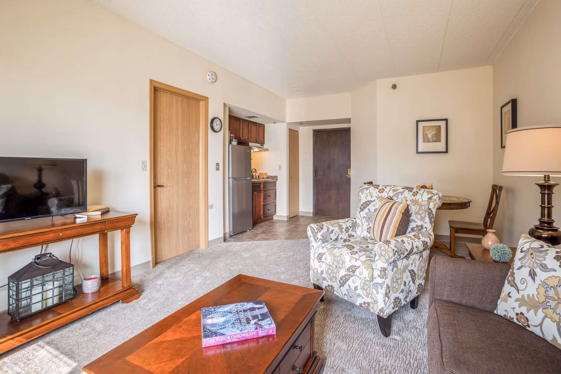 A cozy living room area in a senior living facility with a floral patterned armchair, a brown sofa with matching floral cushions, a wooden coffee table with a book on it, a wooden TV stand with a flat-screen TV, and a small kitchen area with a refrigerator and cabinets in the background. The room has beige walls, carpeted flooring, and a wooden door leading to another room.