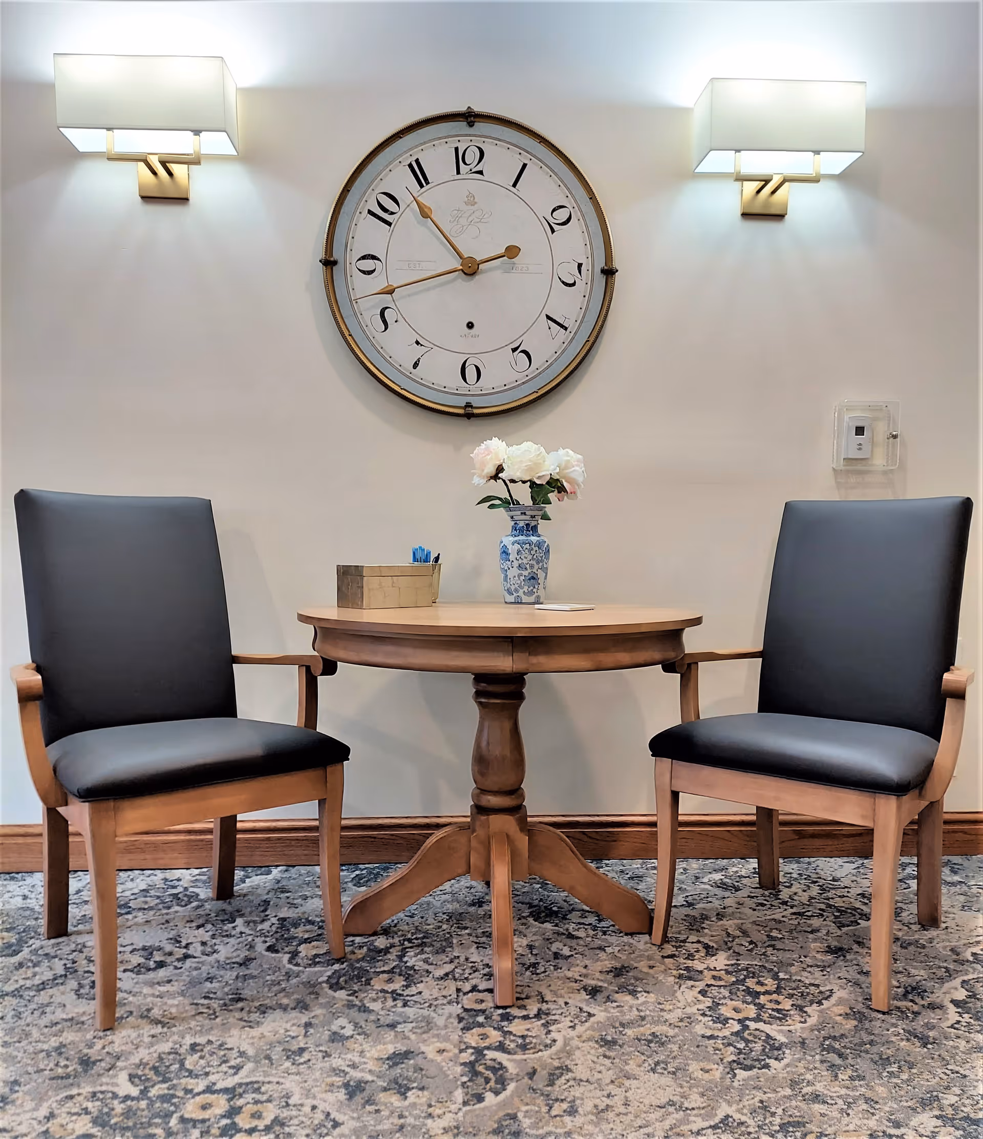 A small round wooden table with a blue and white vase holding white flowers and a small box with pens on top. Two wooden chairs with black cushioned seats and backs are placed on either side of the table. A large round wall clock is mounted on the wall above the table, flanked by two modern wall sconces with white rectangular shades. The floor has a patterned carpet in shades of blue and beige.