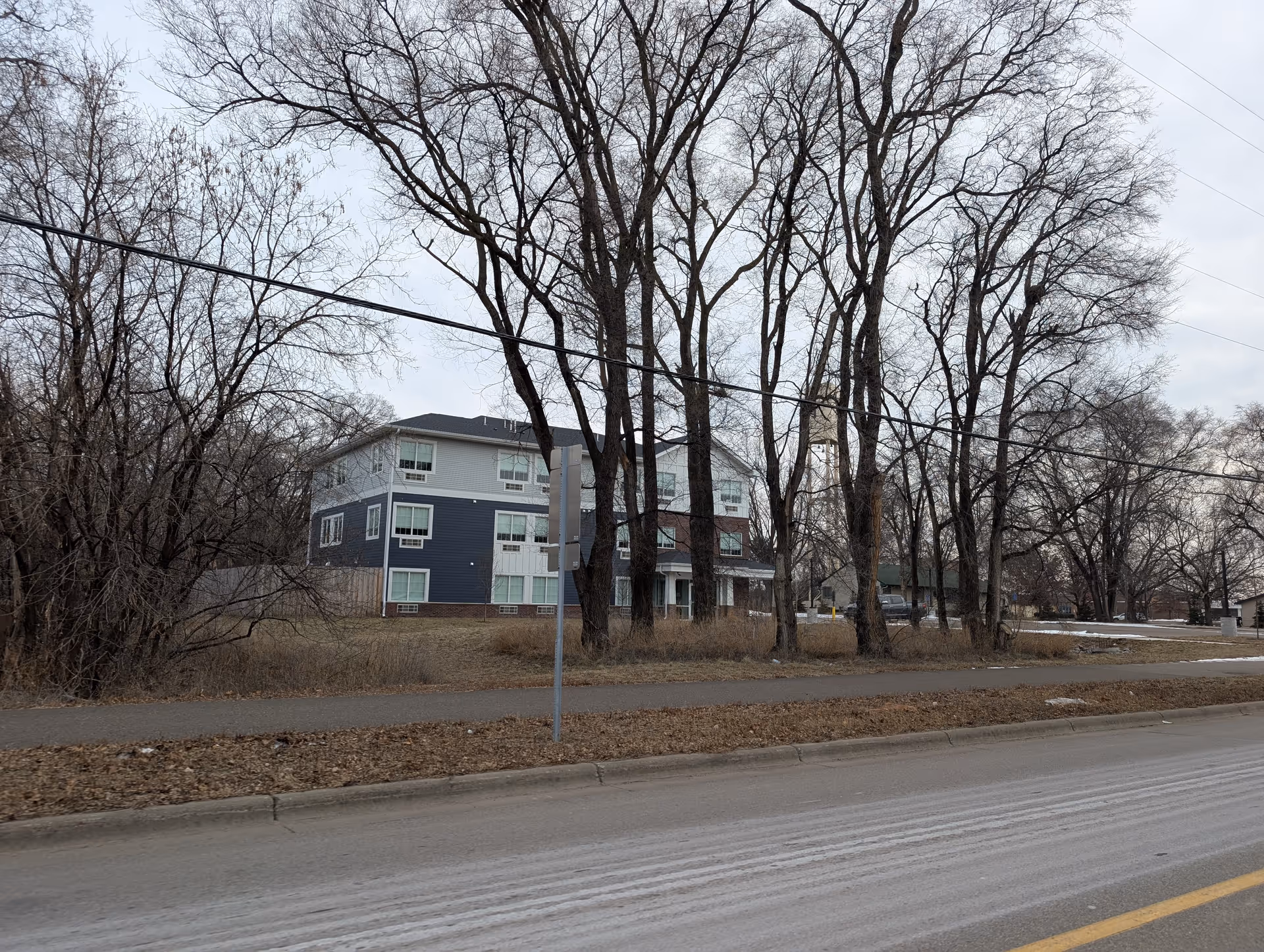 Exterior view of a multi-story residential building partially obscured by leafless trees, with a sidewalk and road in the foreground under an overcast sky.