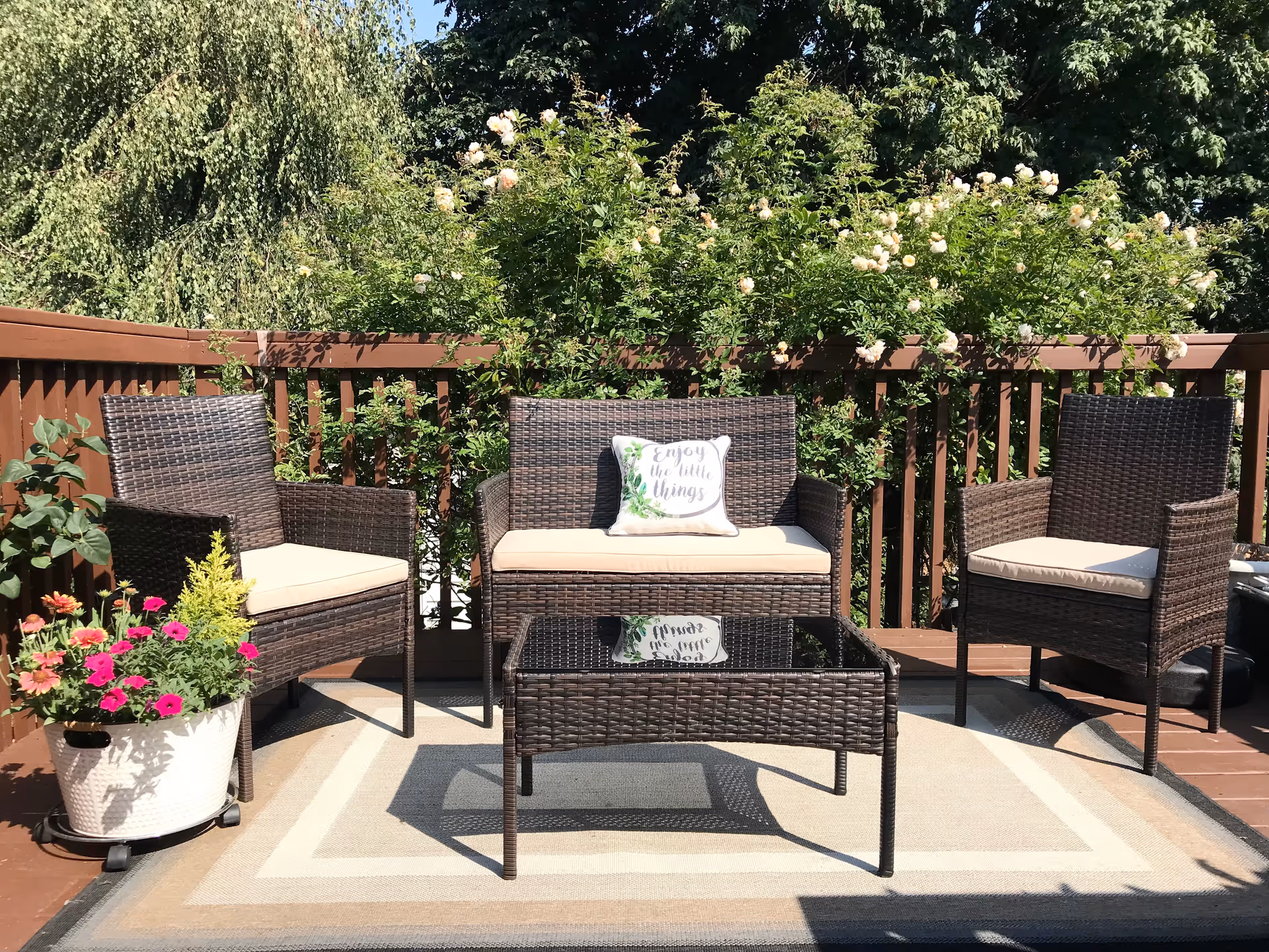 Outdoor patio area with brown wicker furniture including two chairs and a loveseat with beige cushions. A glass-top wicker coffee table sits on a beige and gray outdoor rug. There is a white planter with colorful flowers on the left side and green bushes with white flowers behind the wooden railing.