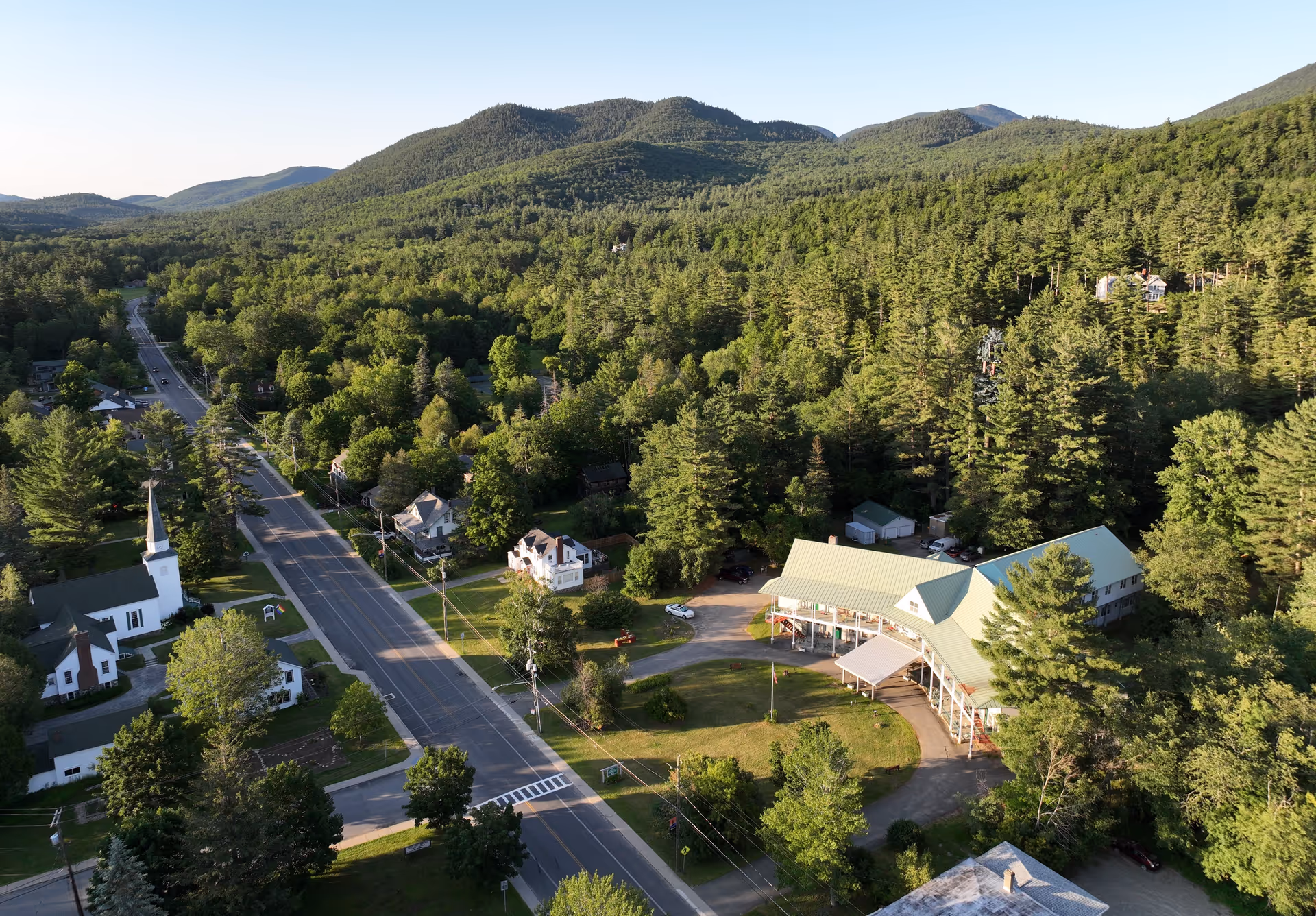 Aerial view of a small town surrounded by dense green forest and hills. A main road runs through the town with houses, a church with a tall steeple, and a large building with a green roof, likely the Keene Valley Neighborhood House. The scene is bathed in natural daylight with clear skies.