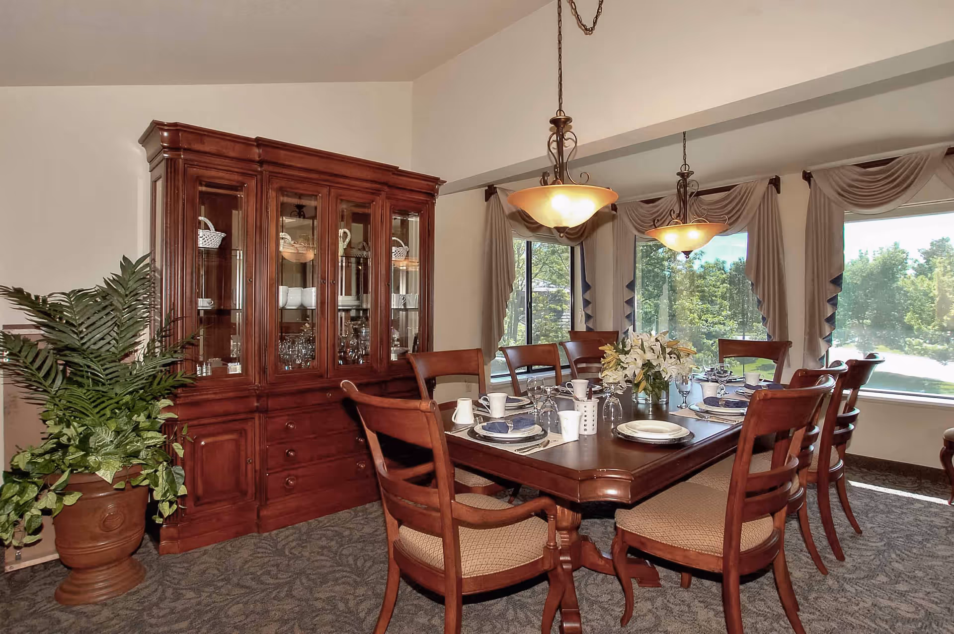 A formal dining room with a large wooden dining table set for eight people, featuring plates, cups, and a floral centerpiece. The room has large windows with draped curtains allowing natural light to enter. A wooden china cabinet filled with dishes and glassware is against one wall, and a potted plant is placed beside it. Two hanging pendant lights illuminate the table.