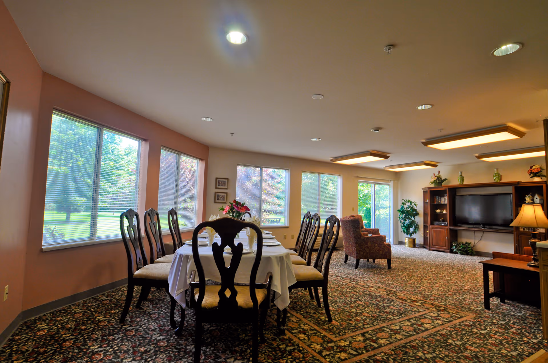 A spacious dining and living area in a senior living facility with a long dining table covered with a white tablecloth and surrounded by wooden chairs. Large windows along the wall let in natural light and offer views of green trees outside. The living area features armchairs, a large TV on a wooden entertainment center, and decorative plants and lamps.