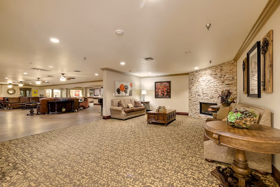 A spacious and warmly lit living room area in a senior living facility with patterned carpet, beige walls, and a stone fireplace. The room features comfortable sofas, a wooden coffee table, decorative wall art, a side table with a floral arrangement, and ceiling fans. In the background, there is an open area with additional seating and tables.