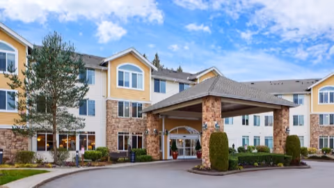 Three-story yellow and beige senior living building with a covered porte-cochère entrance and driveway under a partly cloudy sky.