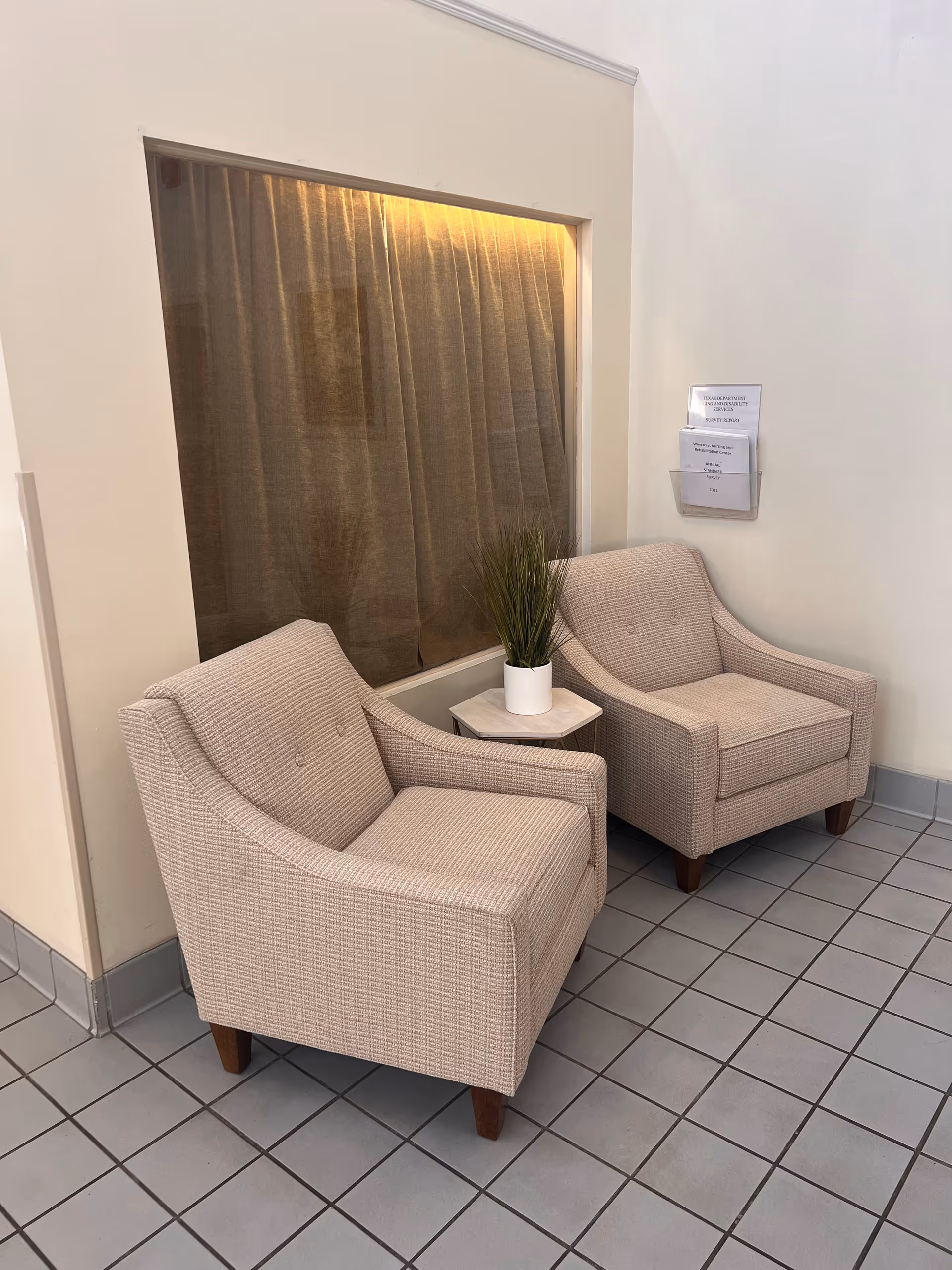 Two beige upholstered armchairs with wooden legs are placed on a tiled floor next to a small hexagonal side table with a white pot containing a green plant. Behind the chairs is a large window with brown curtains and a wall-mounted brochure holder containing papers.