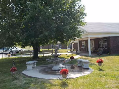 Outdoor courtyard with a stone fountain, red hanging baskets, and a brick senior living building.