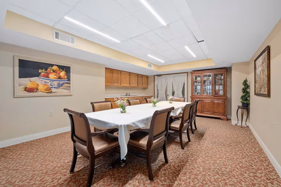 A dining room with a long rectangular table covered with a white tablecloth and surrounded by eight wooden chairs with cushioned seats. The room has beige walls, patterned carpet, a wooden cabinet with glass doors, and two framed paintings on the walls. There are also small flower arrangements on the table and a small plant on a stand in the corner.
