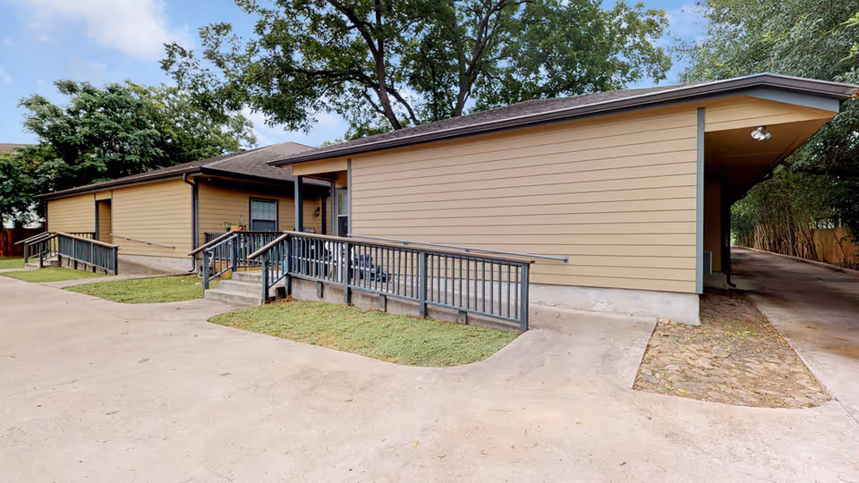 Exterior view of a single-story beige building with a sloped roof, featuring wheelchair ramps and handrails leading to entrances. The building is surrounded by concrete pavement and some grass patches, with trees in the background under a partly cloudy sky.
