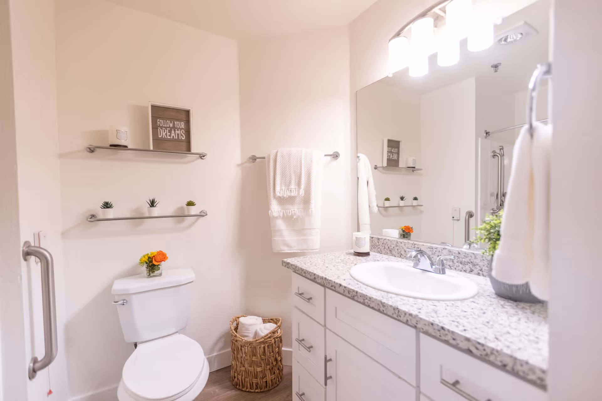 Bright and clean bathroom with a white toilet, granite countertop with a sink, a large mirror, and white cabinets. There are two metal shelves on the wall above the toilet holding small potted plants and a framed sign that says 'FOLLOW YOUR DREAMS'. A white towel hangs on a towel rack next to the sink, and a wicker basket with rolled towels is on the floor beside the toilet. The bathroom has light-colored walls and wood-style flooring.