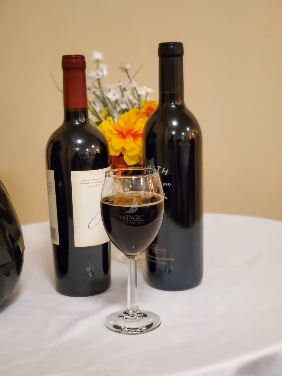 A close-up of a wine glass filled with red wine placed on a white tablecloth. Behind the glass, there are two bottles of red wine and a small bouquet of yellow and white flowers against a beige wall.