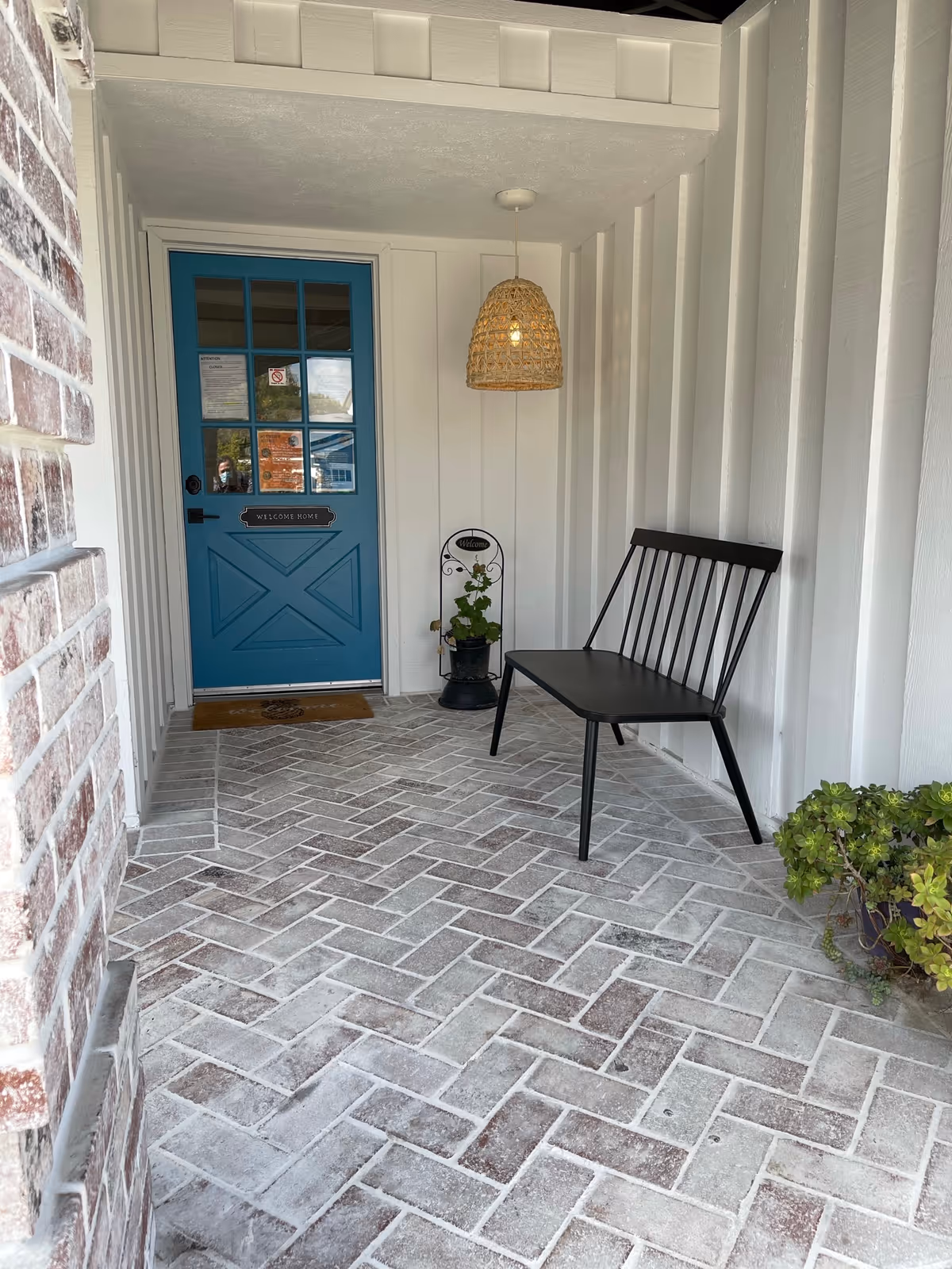 A covered porch area with a blue door featuring glass panes and a 'Welcome Home' sign. The porch has white vertical paneling walls, a woven hanging light fixture, a black metal bench, a small potted plant on a stand, and another potted plant on the floor. The floor is made of light-colored brick arranged in a herringbone pattern.