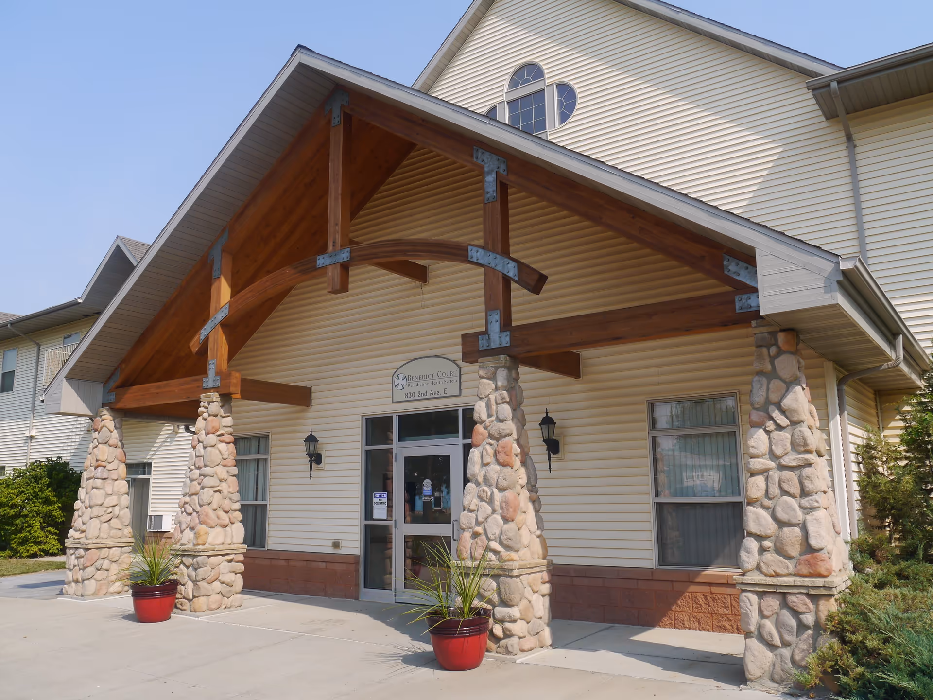 Entrance of Benedict Court facility featuring a covered porch with wooden beams supported by stone pillars, glass double doors, and two potted plants on either side of the entrance.