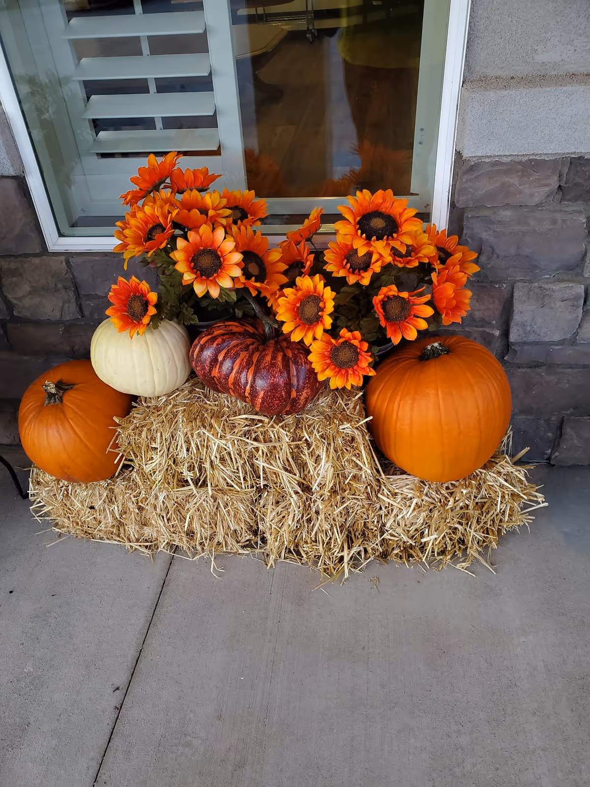A fall display of pumpkins, a hay bale, and bright orange sunflowers arranged in front of a window on a stone exterior.