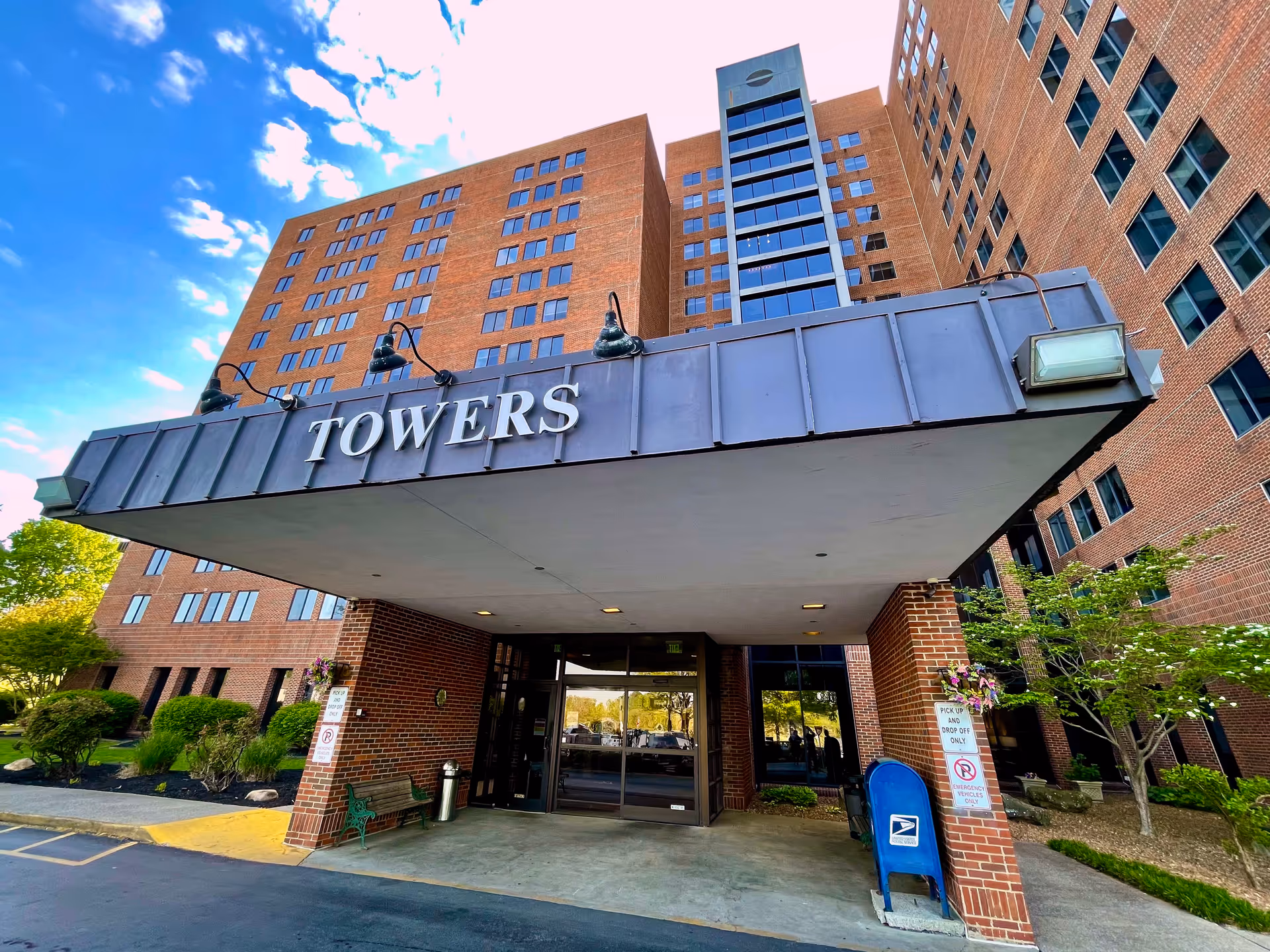 Entrance of a tall brick building with a large overhang displaying the word 'TOWERS'. The building has many windows and a glass elevator shaft in the center. There are some bushes and trees around the entrance, a bench, a mailbox, and a sign indicating pick up and drop off only.