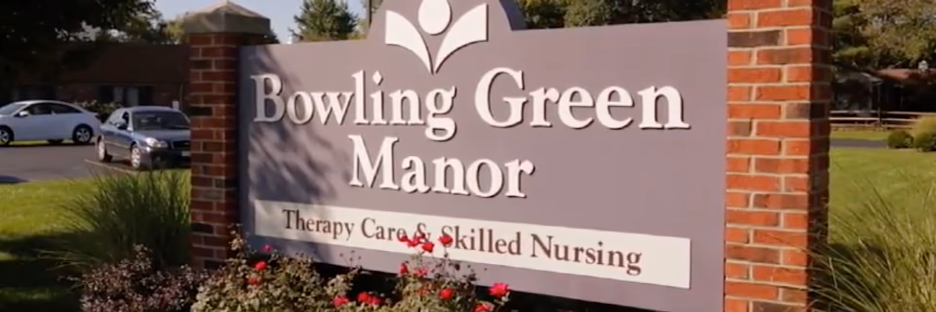 Outdoor sign for Bowling Green Manor, a therapy care and skilled nursing facility, mounted on a brick structure with grass, flowers, and parked cars in the background.