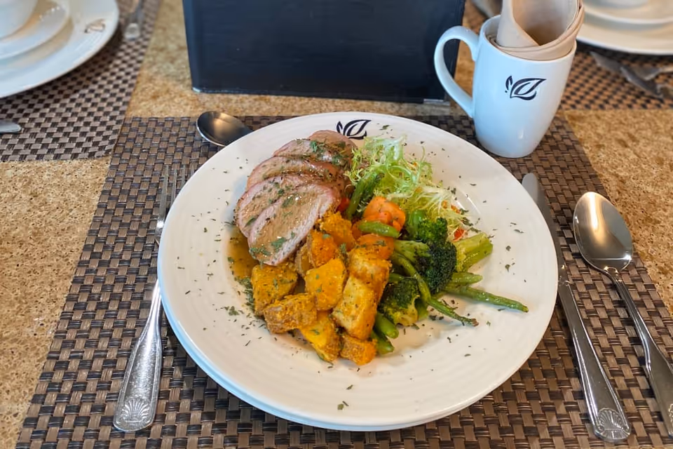 A white plate with sliced meat, roasted vegetables and salad on a placemat, with cutlery and a mug at a dining table.
