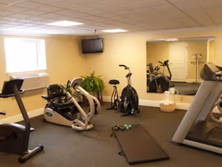 A small exercise room with various fitness equipment including a recumbent bike, an upright stationary bike, a treadmill, and a yoga mat with dumbbells. There is a large mirror on one wall, a flat-screen TV mounted in the corner, a window with an air conditioning unit below it, and a potted plant near the corner.