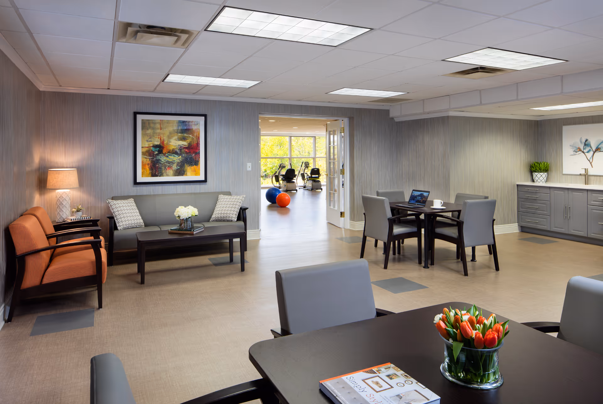 A well-lit common area in a senior living facility featuring a seating area with a gray sofa, two orange armchairs, and a coffee table with a flower arrangement. There are two tables with chairs, one with a laptop and a cup. The room has gray walls with framed artwork, a lamp on a side table, and a cabinet with a plant and artwork above it. In the background, a fitness room with exercise equipment and colorful exercise balls is visible through an open doorway.