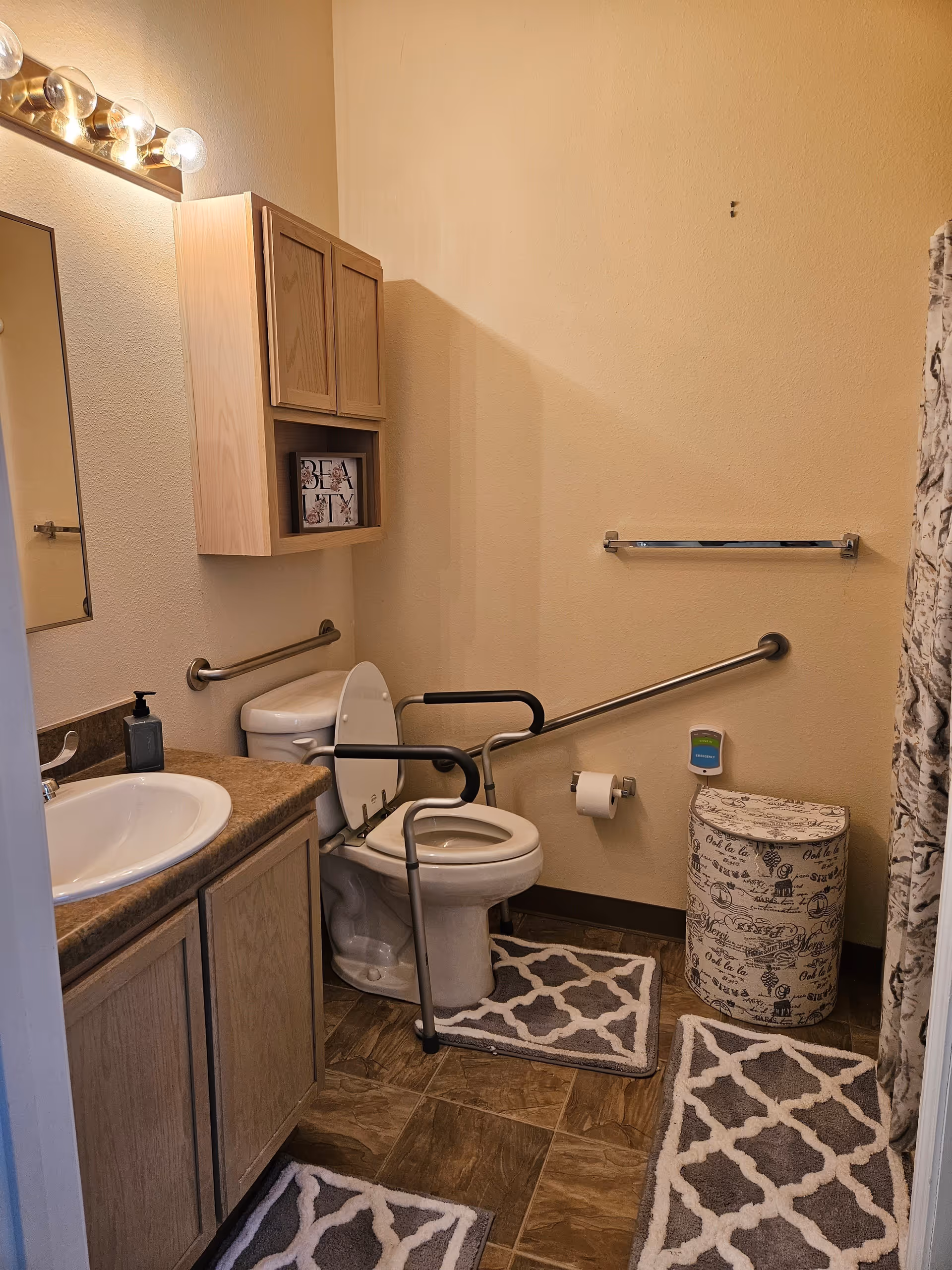A bathroom with a toilet equipped with safety rails, a sink with a countertop and cabinet, a wall-mounted cabinet above the toilet, a towel rack, a decorative laundry hamper, and patterned floor mats. The walls are beige and the floor has brown tiles.