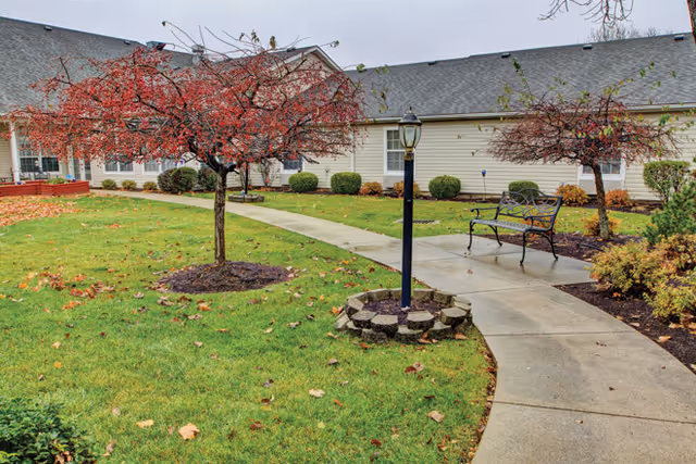A landscaped outdoor area with a curved concrete walkway, green grass, and two small trees with red leaves. There is a black metal bench and a black lamp post surrounded by a circular arrangement of stones. The background shows a single-story beige building with white-framed windows.