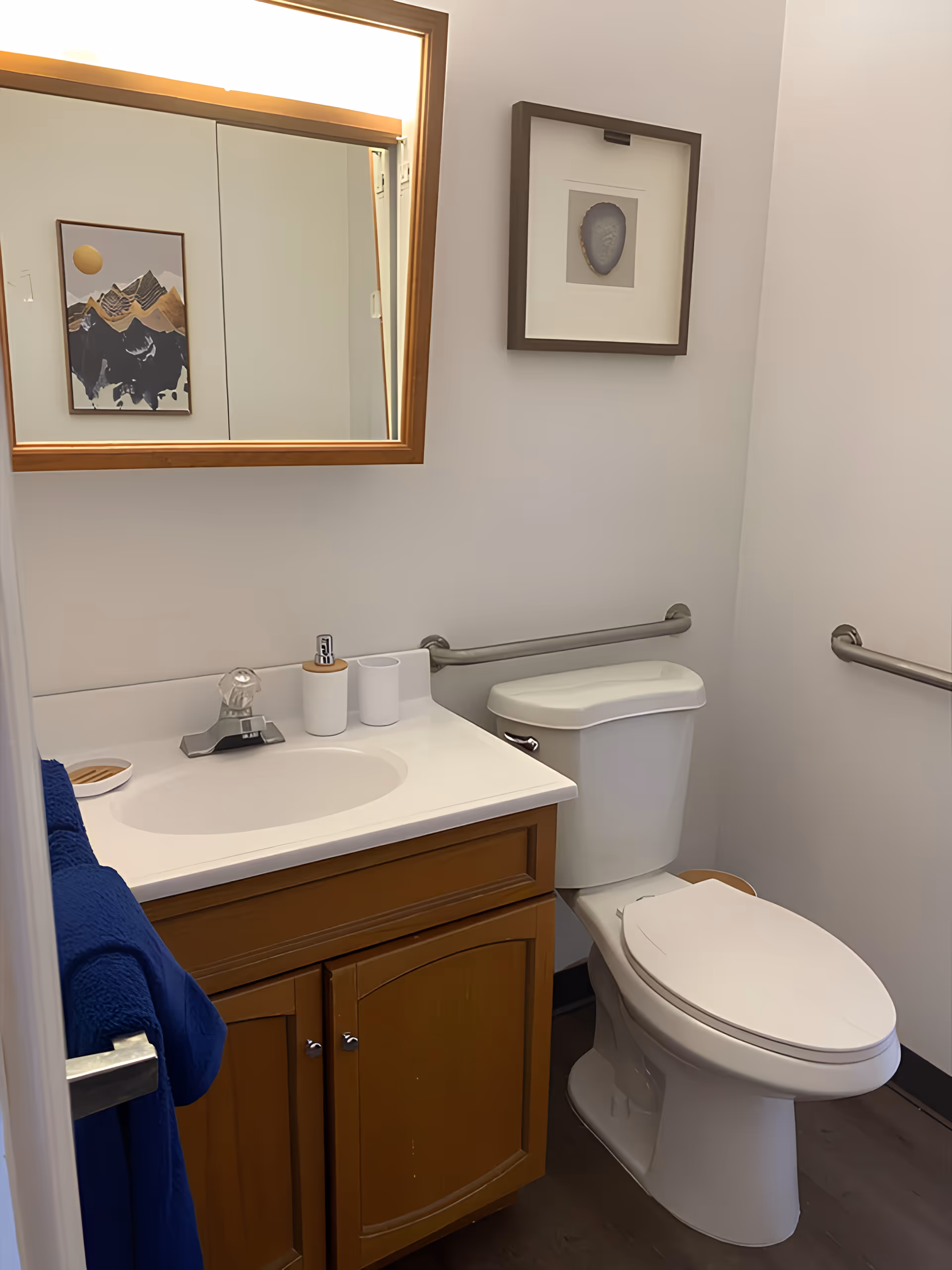 A small bathroom with a white toilet and a wooden vanity with a white sink. Above the sink is a mirror with a wooden frame and a light fixture. There are two framed pictures on the walls, one reflected in the mirror and one above the toilet. A blue towel hangs on a metal towel bar next to the vanity. The bathroom has grab bars on the walls near the toilet.