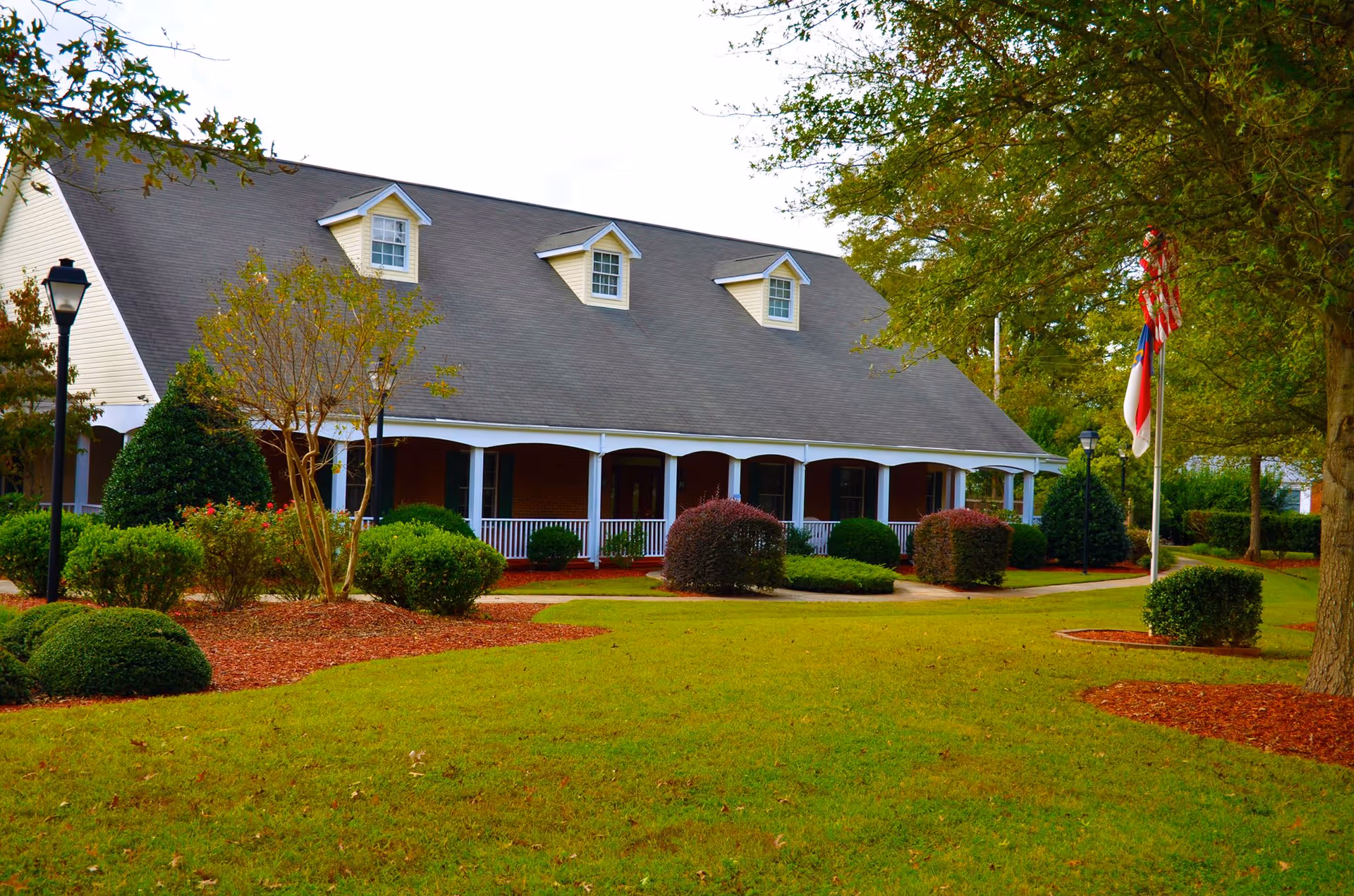 Front exterior view of a single-story assisted living facility building with a large porch, surrounded by well-maintained green lawn, bushes, trees, and a flagpole with flags.