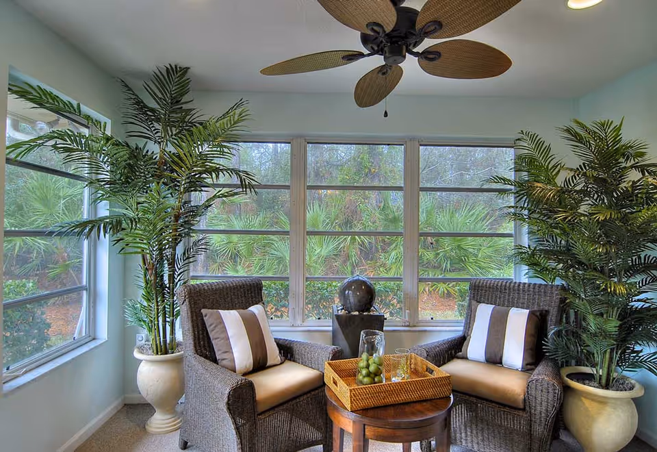 A cozy sunroom with two wicker armchairs featuring beige cushions and striped pillows, a small round wooden table with a wicker tray holding a glass pitcher and glasses, large potted plants on either side, a ceiling fan with leaf-shaped blades, and large windows showing greenery outside.