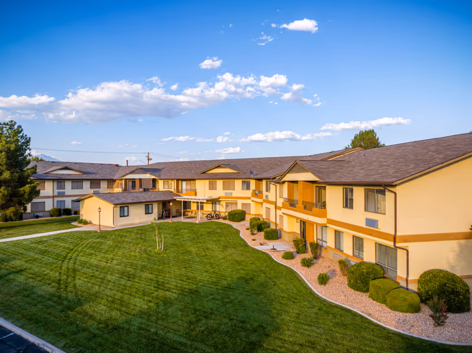 Exterior view of a two-story senior living facility building with beige walls and brown roof, surrounded by well-maintained green lawn and shrubs under a blue sky with scattered clouds.