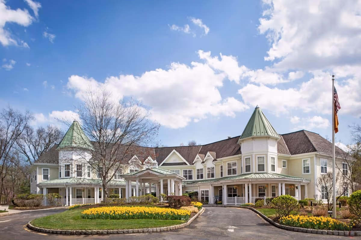 Front exterior of a large two-story cream-colored senior living building with green turrets, a covered driveway entrance, flagpole, and landscaped flower beds under a blue sky.