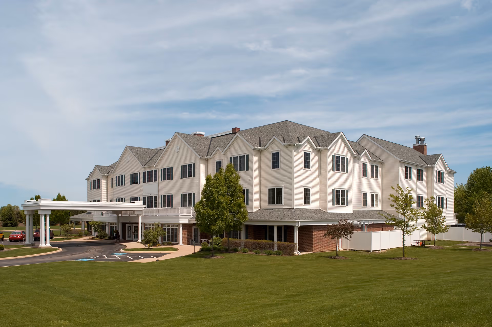 Exterior view of a large, three-story assisted living community building with beige siding and multiple windows. The building has a covered entrance with white columns and a driveway leading up to it. There is a well-maintained green lawn and several trees surrounding the building under a partly cloudy sky.