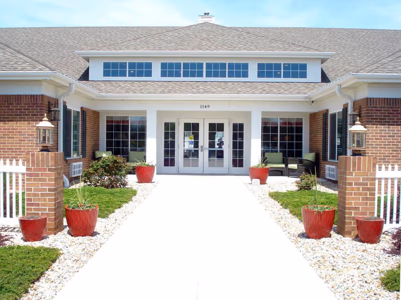Front entrance of a brick senior living building with double glass doors, a paved walkway, and red potted plants.