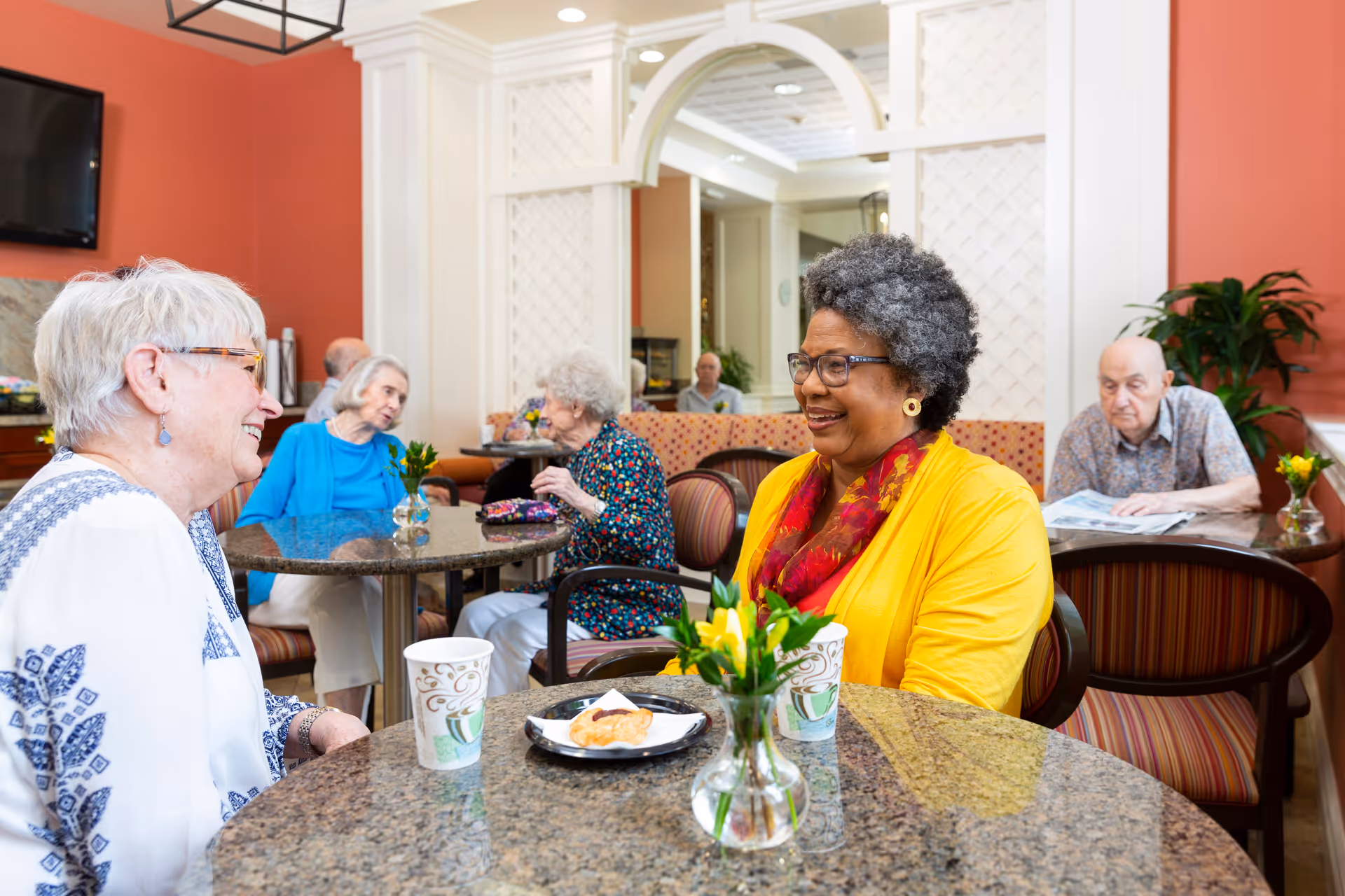 Older adults seated at round tables in a bright communal dining room, talking and enjoying snacks.