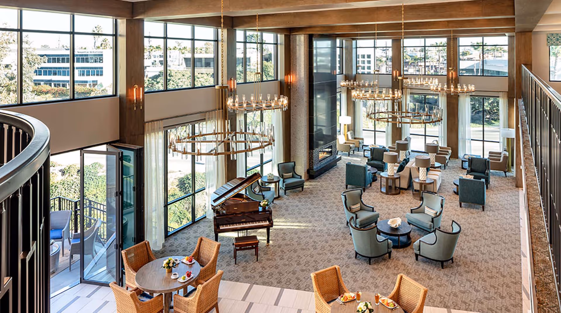 Spacious and elegant senior living facility lounge with large windows allowing natural light, multiple seating areas with armchairs and tables, a grand piano, chandeliers, and an outdoor patio visible through open glass doors.