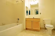 A clean bathroom featuring a bathtub with tiled walls, a wooden vanity with a sink and mirror above it, and a white toilet. The floor has a light-colored tile pattern and the walls are painted a soft yellow.