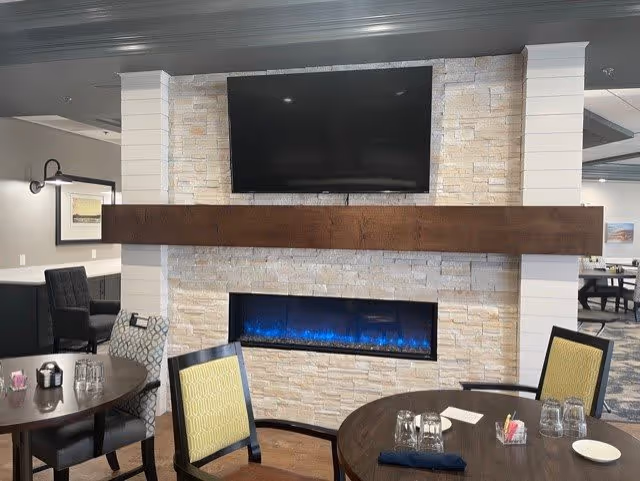 Interior view of a senior living facility dining area featuring a modern electric fireplace with blue flames set in a stone wall. Above the fireplace is a mounted flat-screen TV. The room has round dining tables with chairs, some with yellow patterned upholstery and others with dark upholstery. The background shows additional seating and framed artwork on the walls.