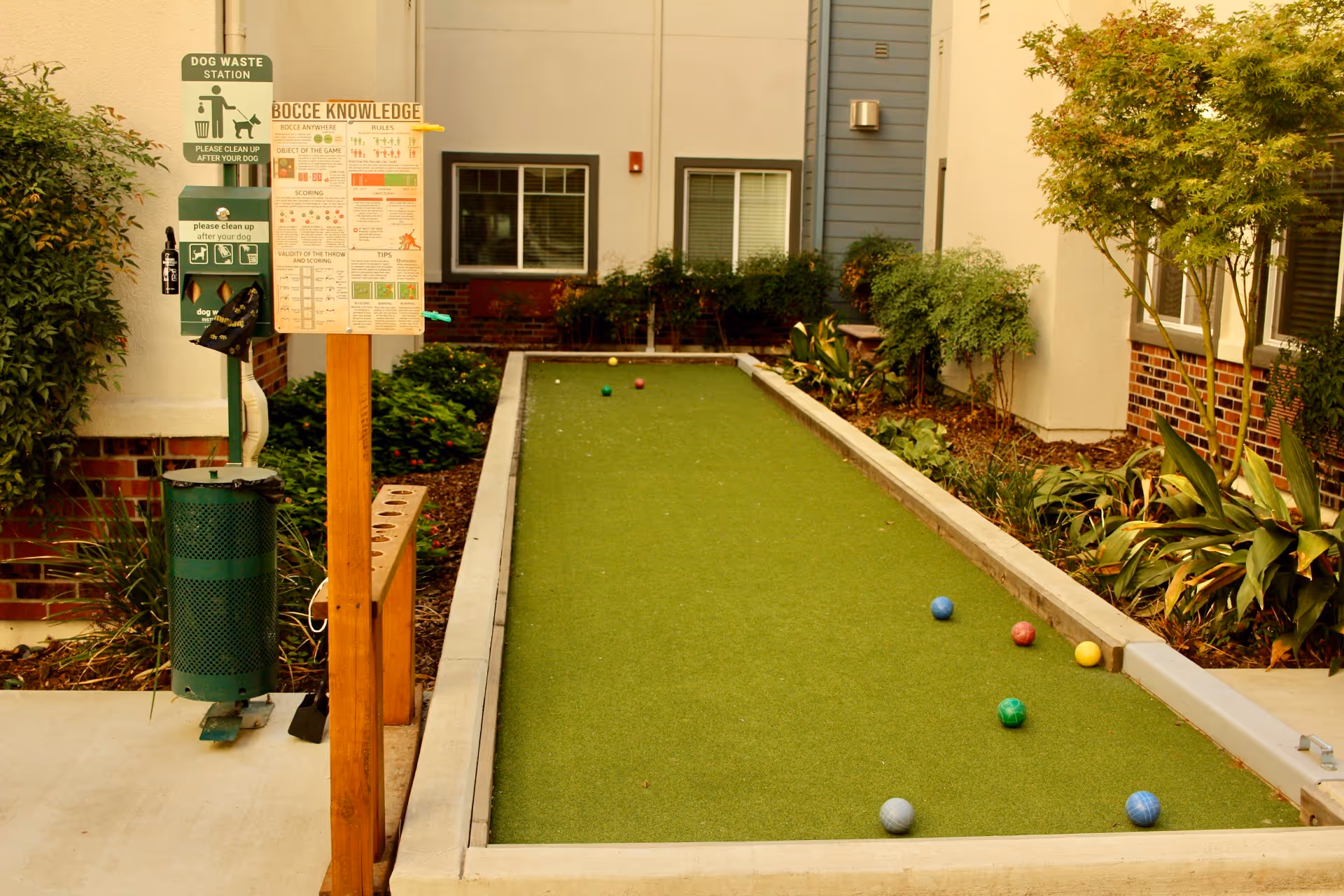 Outdoor bocce ball court in a landscaped courtyard with scattered bocce balls and a nearby dog waste station.