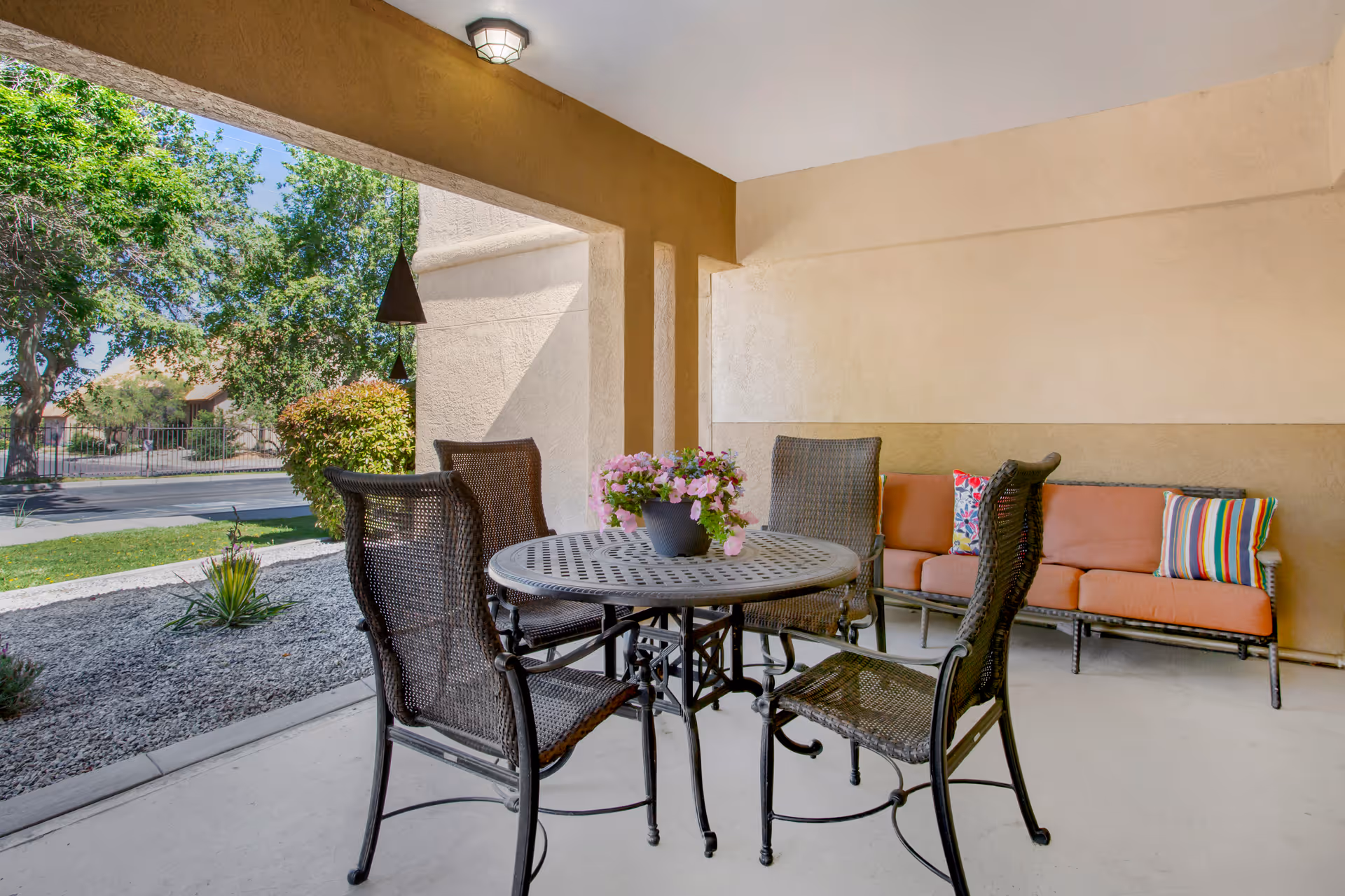 Covered outdoor patio area with a round metal table and four wicker chairs surrounding it. A potted plant with pink flowers is on the table. In the background, there is a wicker sofa with orange cushions and colorful striped pillows. The patio overlooks a landscaped area with gravel, plants, and trees, and a street is visible beyond the greenery.