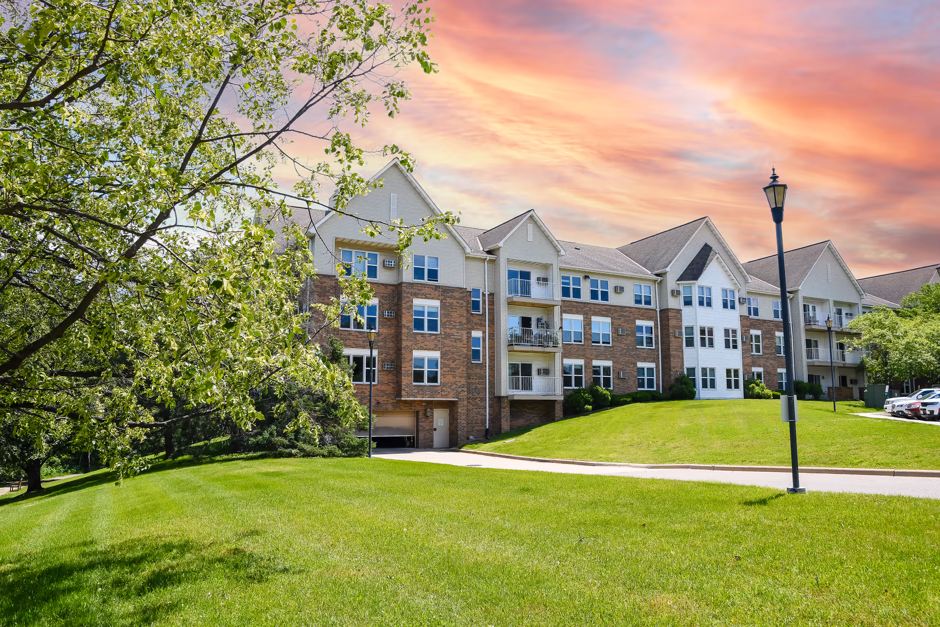 Front exterior of a multi-story senior living apartment building with balconies, a manicured lawn, trees, and a lamp post under a colorful sunset sky.
