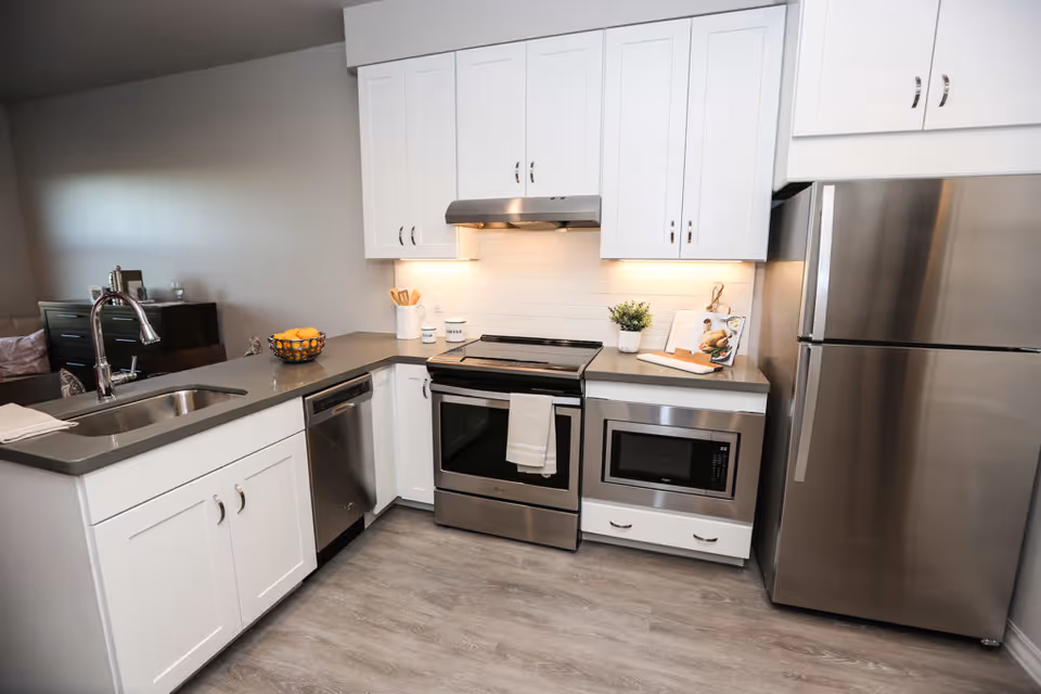 Modern kitchen with white cabinets, stainless steel refrigerator, oven, microwave, dishwasher, and a sink in a gray countertop island.